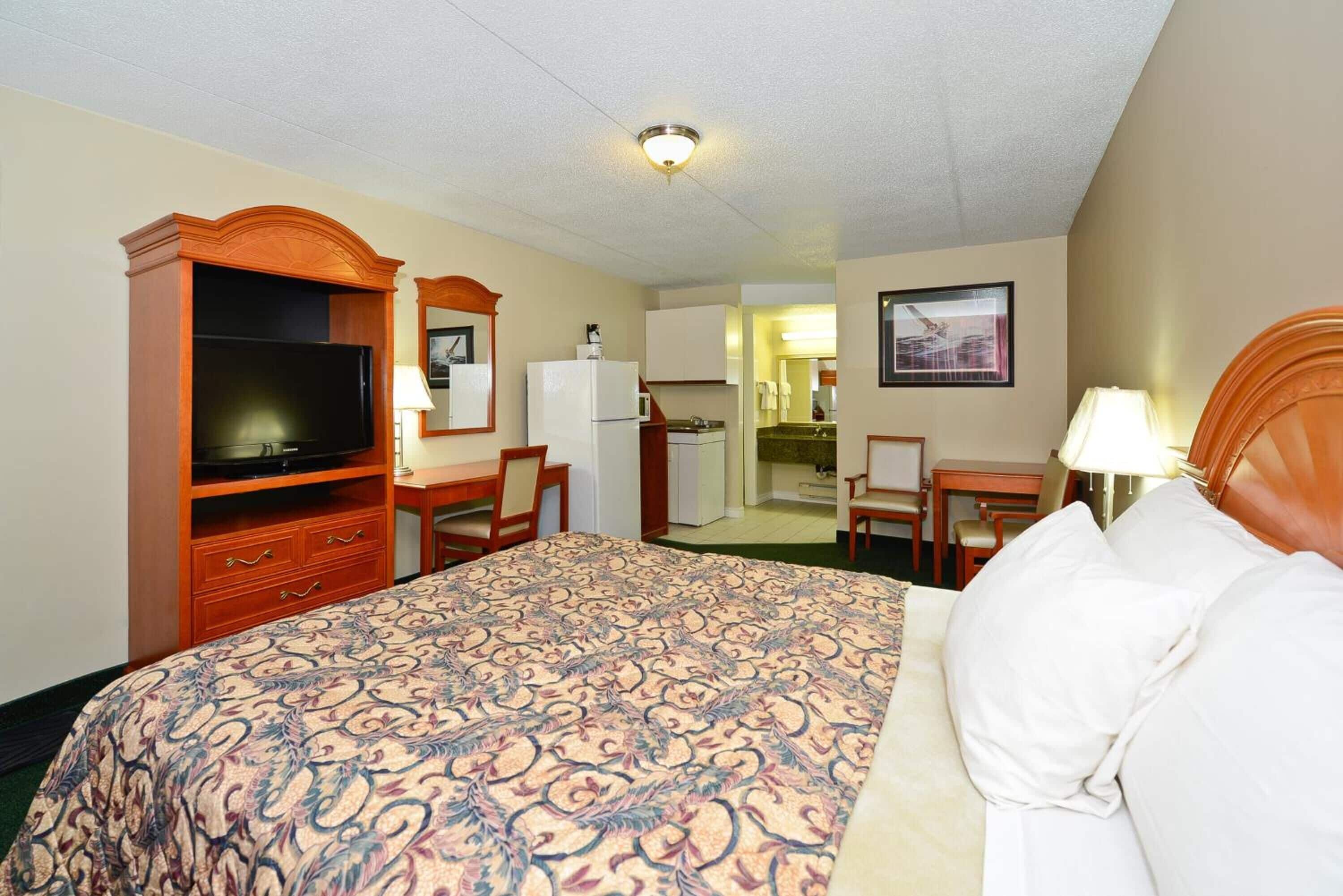 Habitación de hotel con una cama grande, televisor, escritorio con silla, refrigerador y un área de comedor visible a través de una puerta abierta.