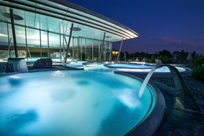A modern outdoor pool area with multiple hot tubs, a glass-enclosed lounge, and a clear view of the surrounding landscape. - Resort Barrière Ribeauvillé (Ribeauvillé)