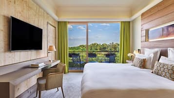 Habitación de hotel con cama, escritorio, silla, televisor y balcón con vistas a zonas verdes.