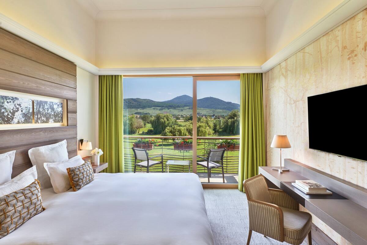 Une chambre d’hôtel avec un grand lit, un bureau, une télévision et un balcon offrant une vue sur la campagne.