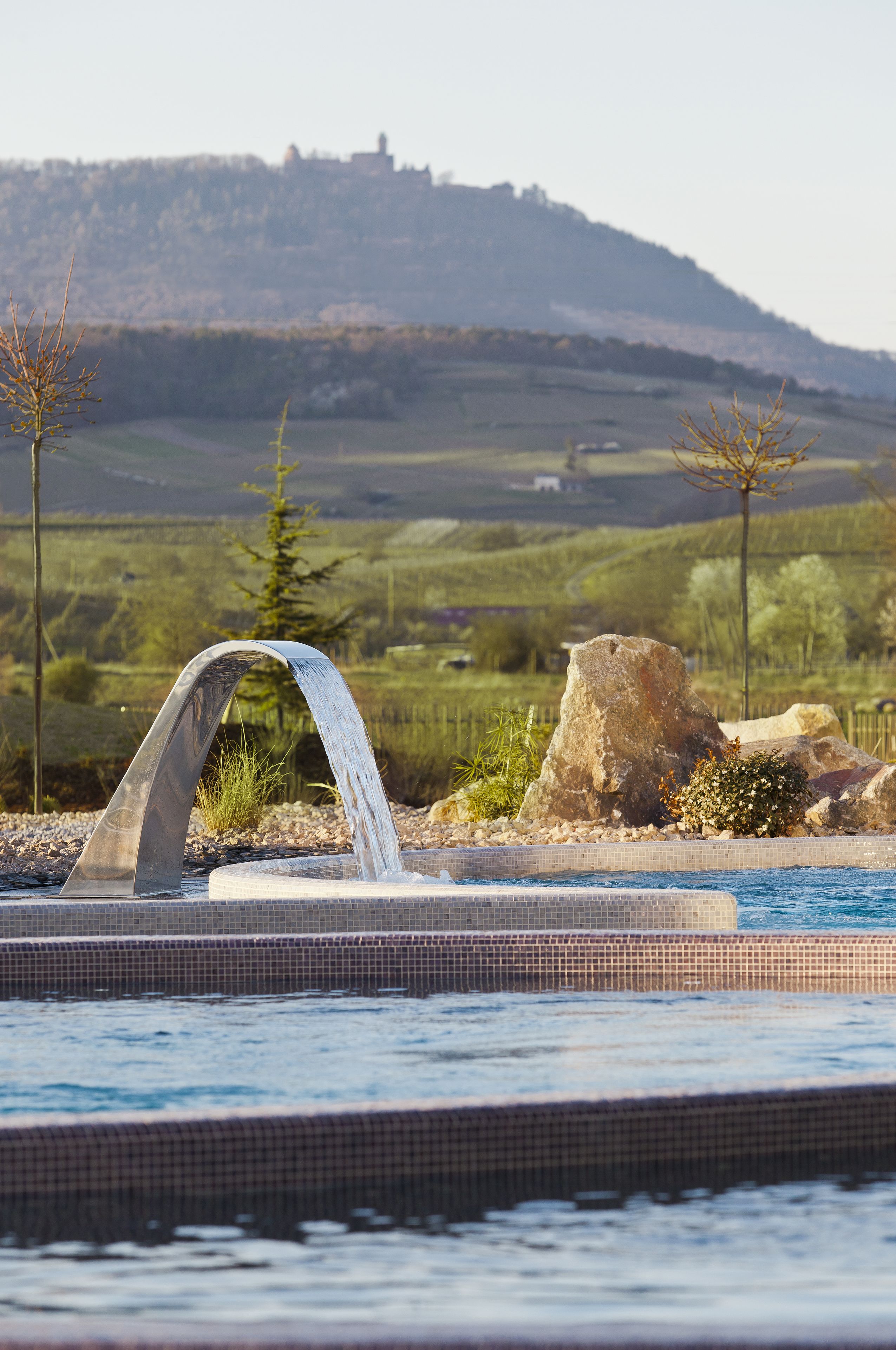 A pool with a waterfall feature, surrounded by rocks and plants, with a hilly landscape in the background.