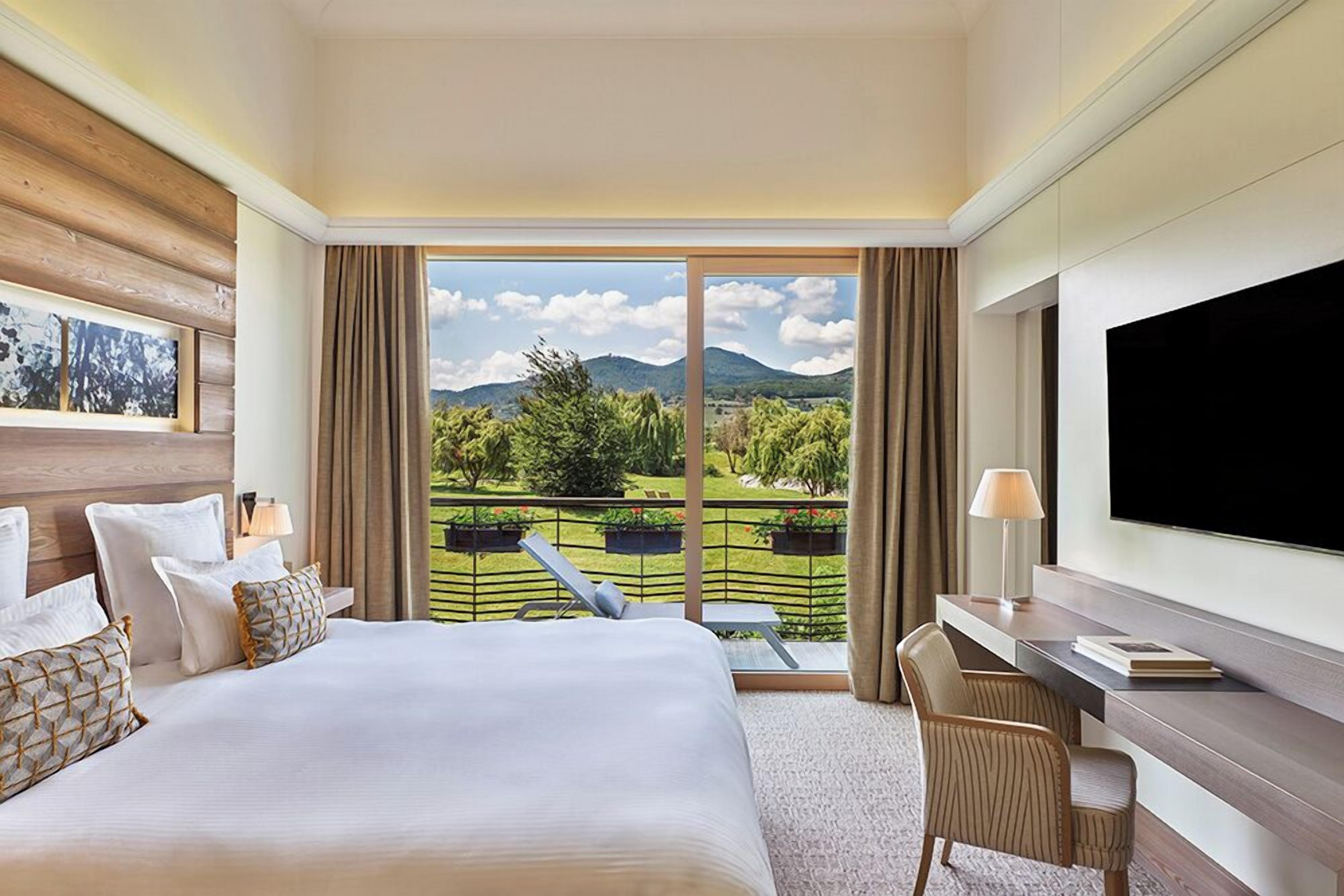 A hotel room with a large bed, a desk, a TV, and a balcony with a view of greenery and mountains.