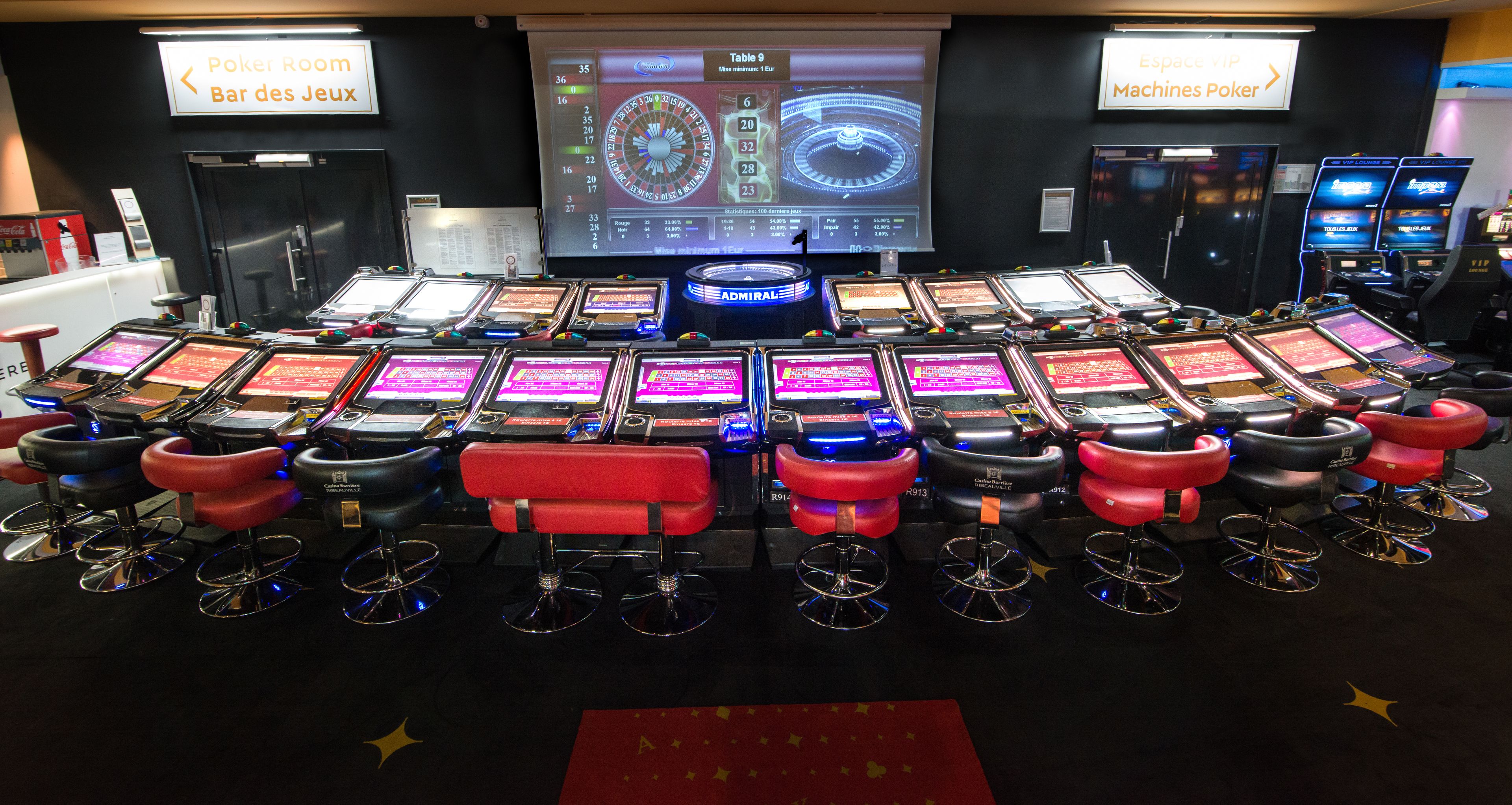 Une salle de poker dotée de plusieurs tables et chaises de jeu, d’un grand écran affichant les statistiques du jeu, et de panneaux indicateurs indiquant la direction vers la « salle de poker » et les « machines à poker ».