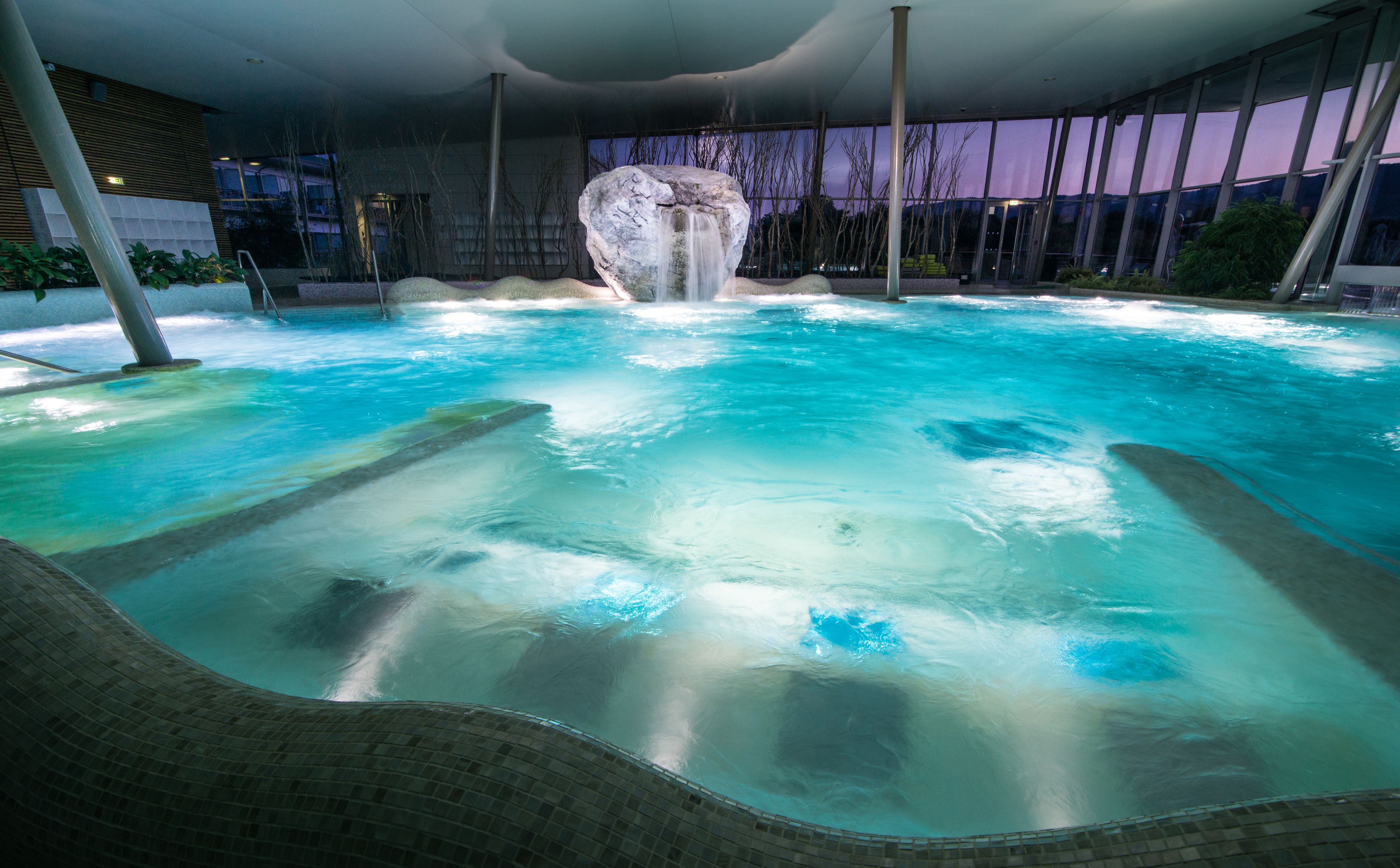 A large indoor swimming pool with a waterfall feature, surrounded by modern architecture and glass walls.