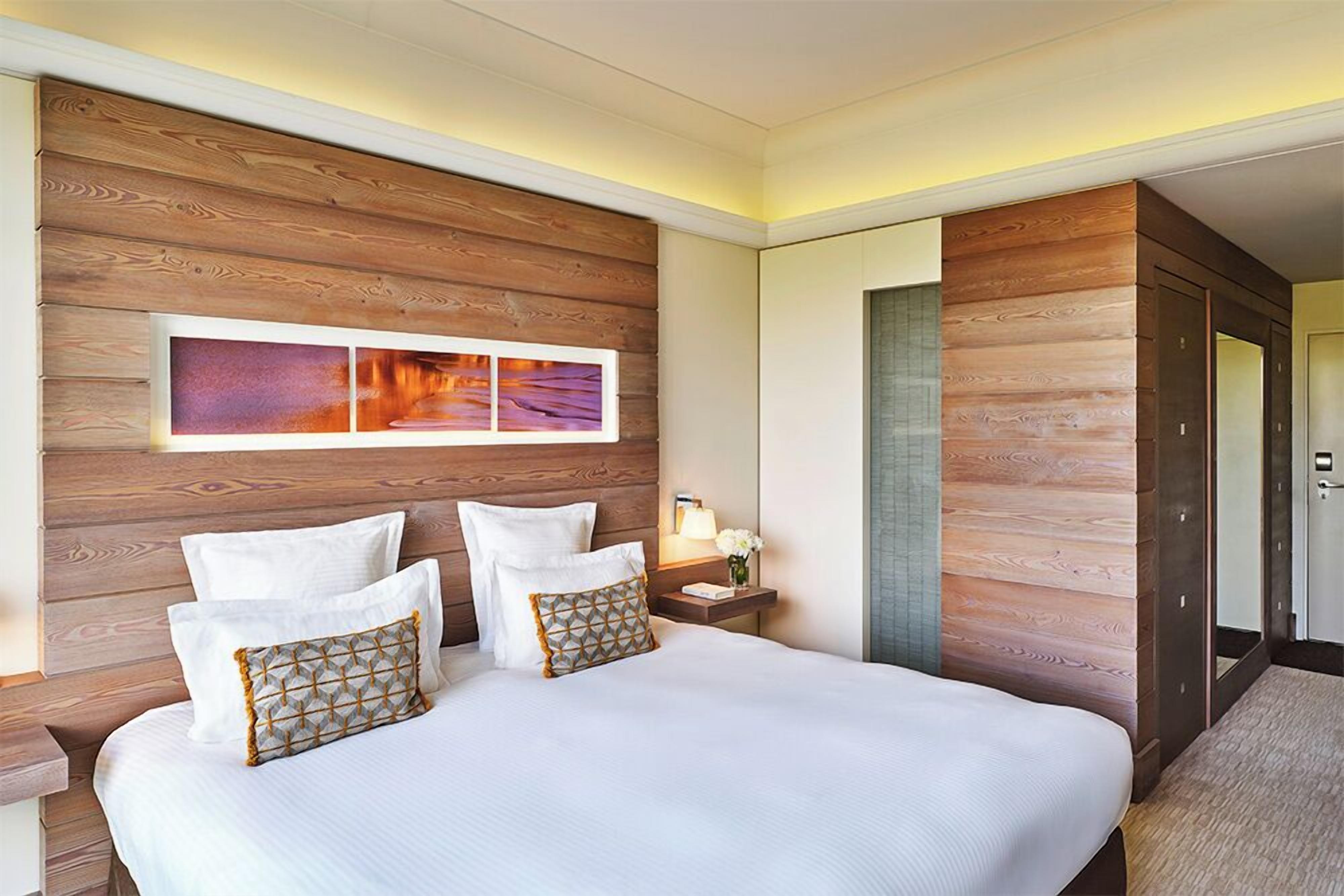 A hotel room with a wooden headboard, a large bed with white bedding, and a nightstand with a lamp and flowers.