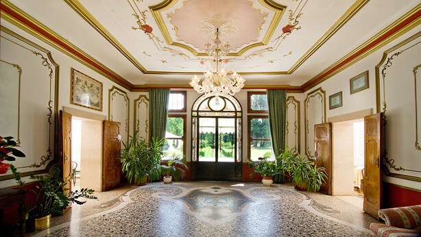 Un amplio pasillo con candelabro, ventanas grandes y elaboradas decoraciones en el techo.