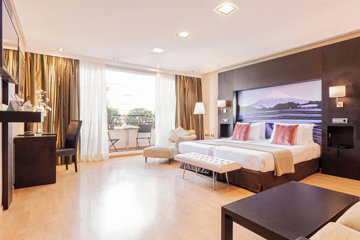 Une chambre d’hôtel avec un grand lit, un téléviseur fixé au mur, un balcon avec une table et des chaises, et un plancher en bois.