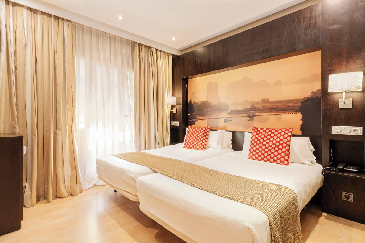 Une chambre d’hôtel avec un grand lit, deux oreillers, une table de chevet avec une lampe, une petite table de nuit avec un téléphone et une grande fenêtre avec des rideaux.