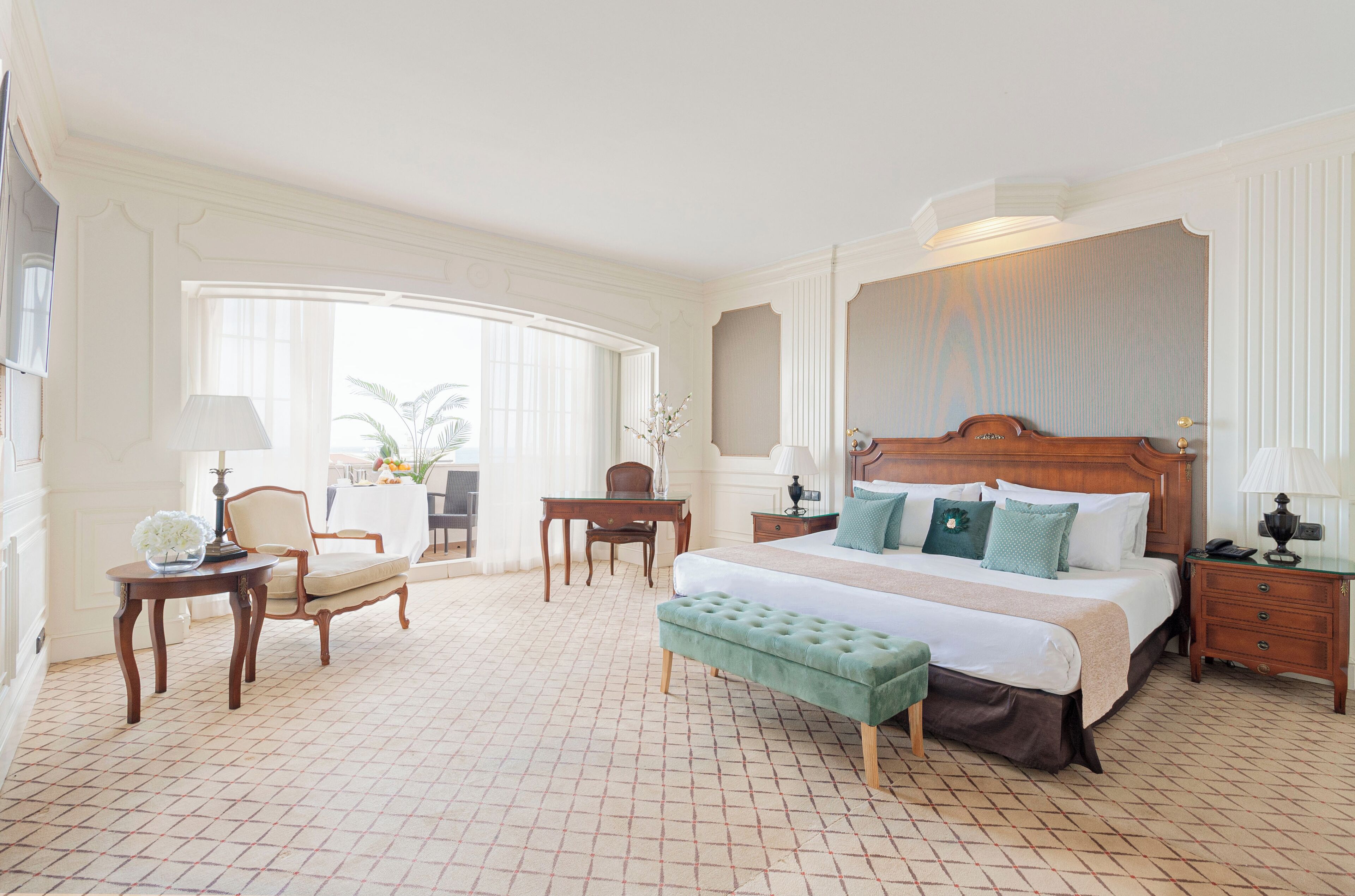 A hotel room with a large bed, a sitting area with a chair and table, a desk, and a small balcony with a view.