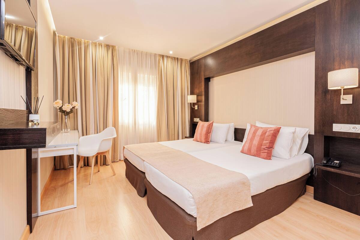 Une chambre d’hôtel avec un grand lit, un bureau et une chaise. Il y a une lampe fixée au mur et un téléphone sur la table de chevet.