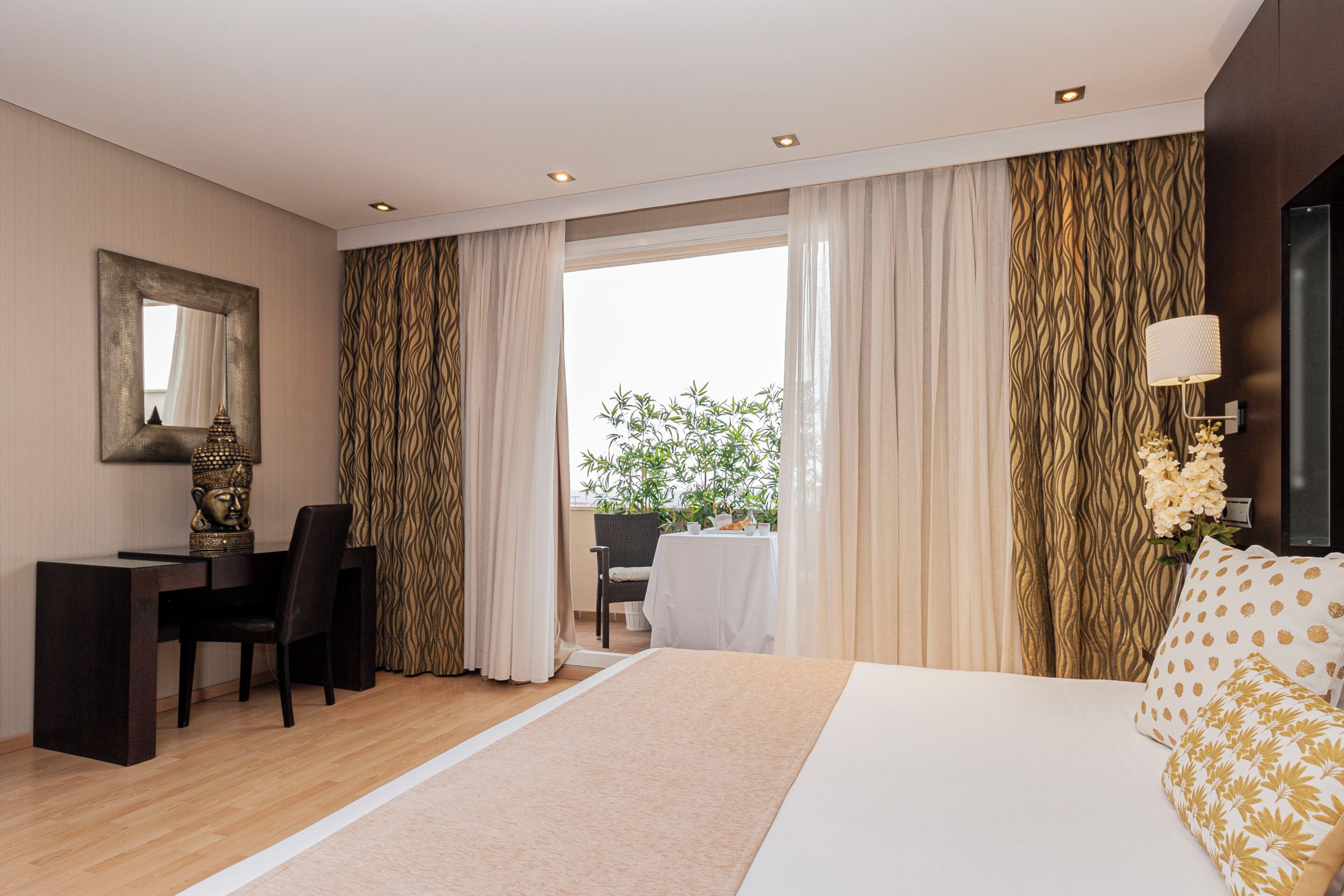 Ein modernes Hotelzimmer mit einem großen Bett, einem Schreibtisch mit Stuhl, einem Essbereich mit Tisch und Stühlen sowie Blick auf Grünfläche.