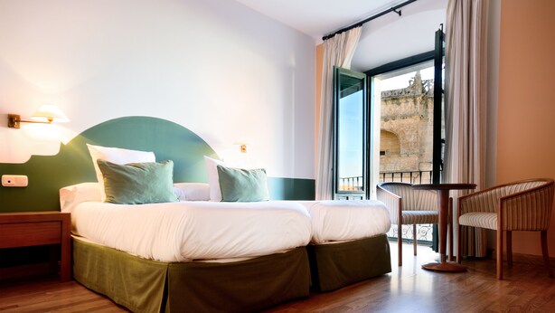 A hotel room with a large bed, a small table, and a view of a historic building through the window. - Hotel AHC Palacio Coria (Coria)