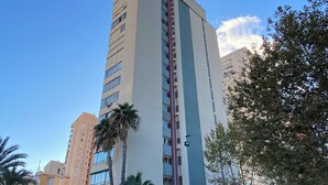 Un grand immeuble à étages, avec une bande verticale rouge au centre, entouré de palmiers et d’autres plantes vertes.