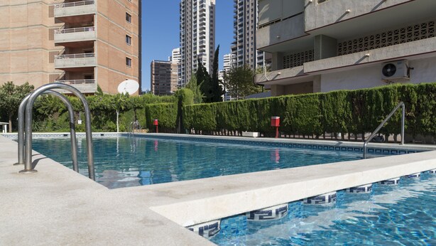 Une piscine à structure métallique, entourée d’une terrasse en béton. Au fond, on aperçoit de grands immeubles d’habitation.