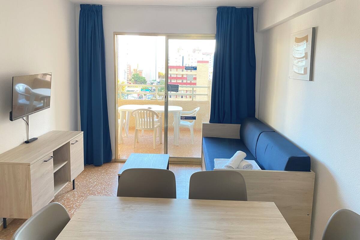 A hotel room with a balcony, a TV, a desk, a sofa, and a dining table.