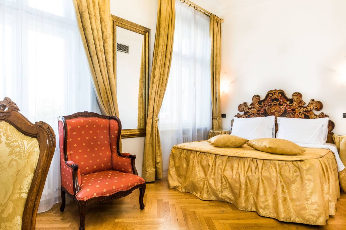 Een hotelkamer met een houten vloer, een rode fauteuil, een bed met een gouden sprei en een grote spiegel aan de muur.