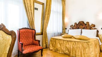 Quarto de hotel com piso de madeira, uma poltrona estofada vermelha, uma cama com colcha dourada e um espelho grande na parede.