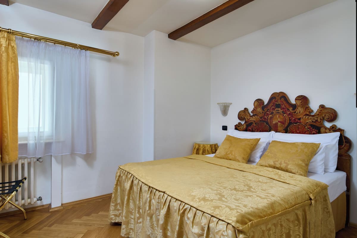 Een slaapkamer met een houten vloer, een bed met een gouden sprei en bijpassende kussens, een houten hoofdbord en een raam met witte gordijnen.