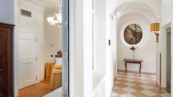Um corredor com piso xadrez, uma janela redonda com grade de metal e um candelabro.