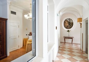 Um corredor com piso xadrez, uma janela redonda com grade de metal e um candelabro.
