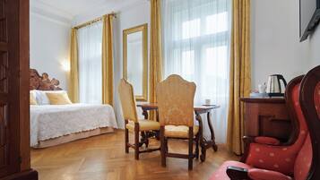 Quarto de hotel com uma cama, uma cadeira de madeira, um poltrona vermelha, uma mesa e um lustre.
