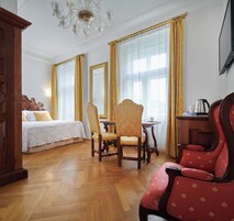 Quarto de hotel com uma cama, uma cadeira de madeira, um poltrona vermelha, uma mesa e um lustre.