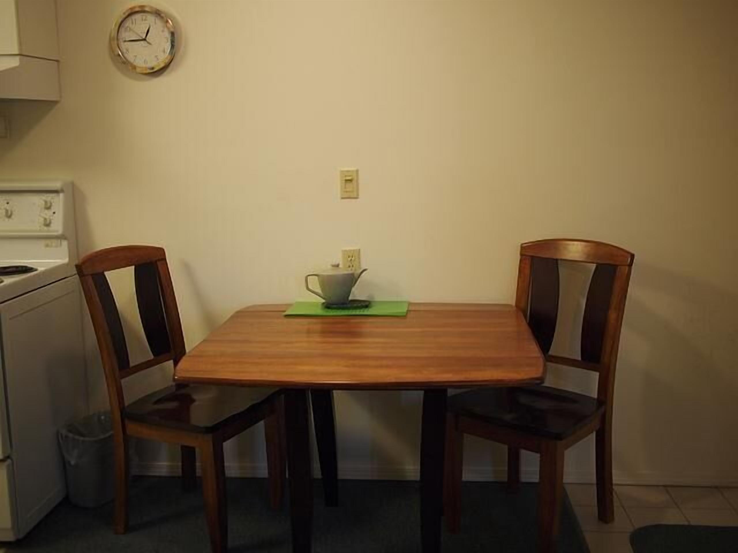 A small, simple dining area with a wooden table and two chairs. A clock is mounted on the wall above the table.