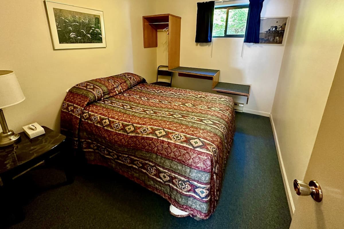 A small, compact room with a bed, a nightstand, a desk, and a window with curtains.