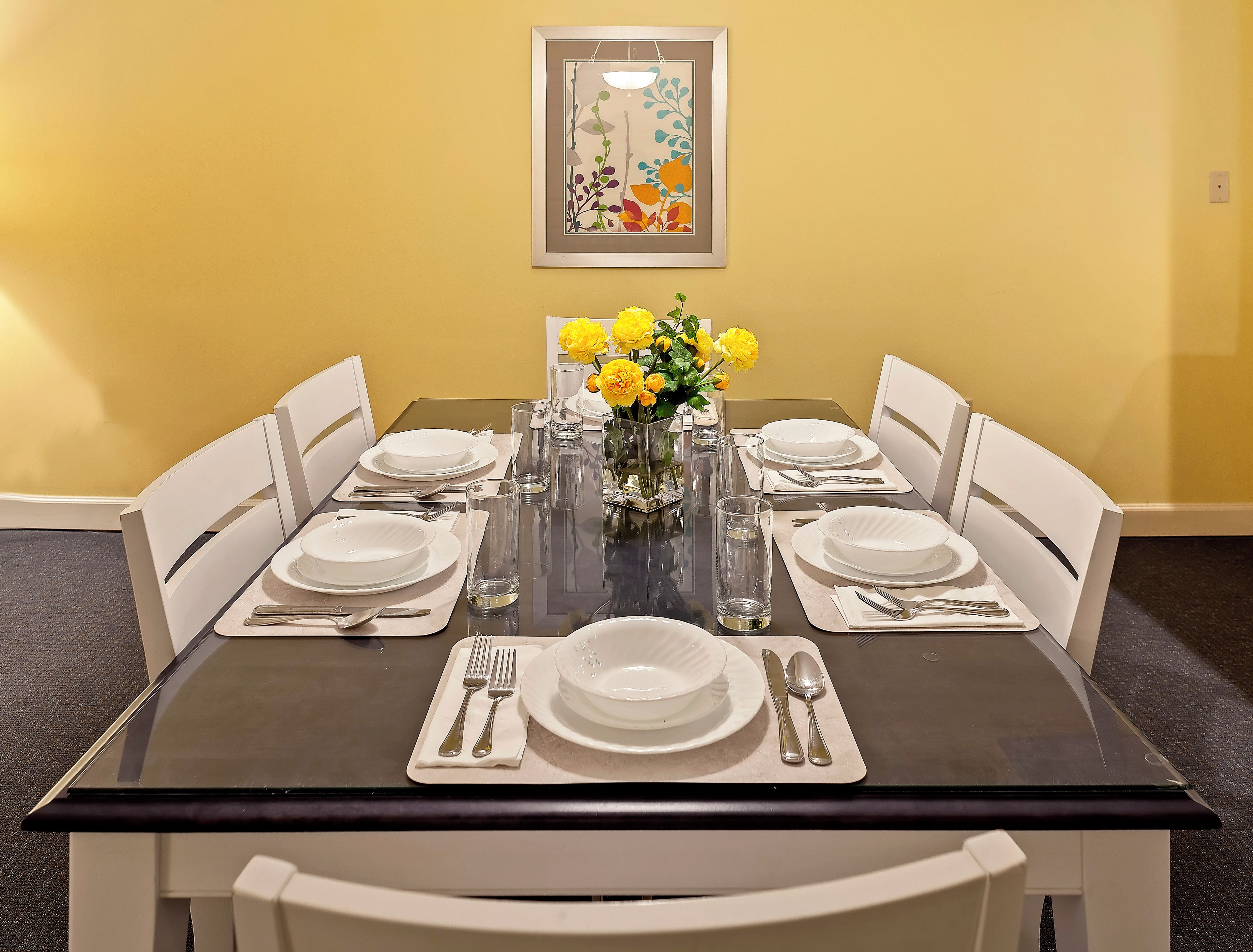 Mesa de comedor preparada para seis personas, con sillas blancas, un jarrón de flores amarillas y un cuadro en la pared.