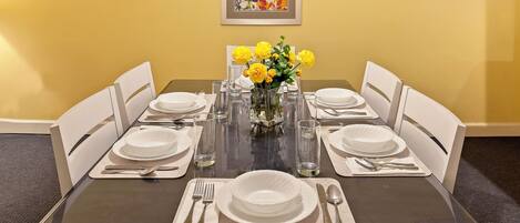 Mesa de comedor preparada para seis personas, con sillas blancas, un jarrón de flores amarillas y un cuadro en la pared.