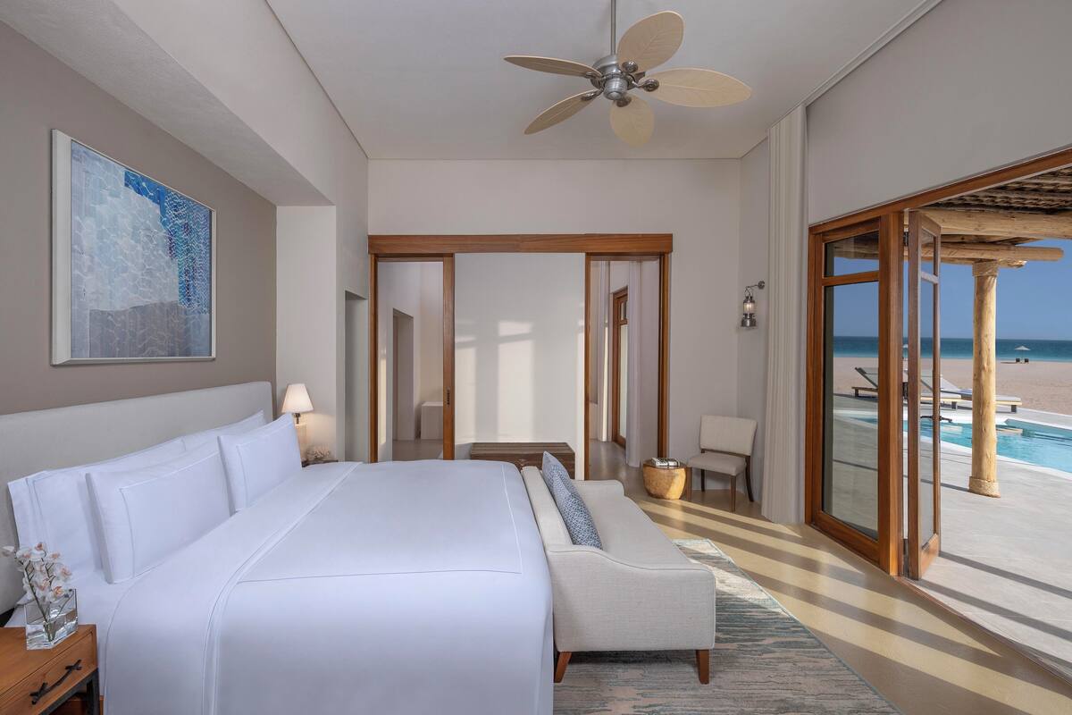 Ein Hotelzimmer mit einem großen Bett, einem Deckenventilator, Blick auf den Strand und einem Pool.
