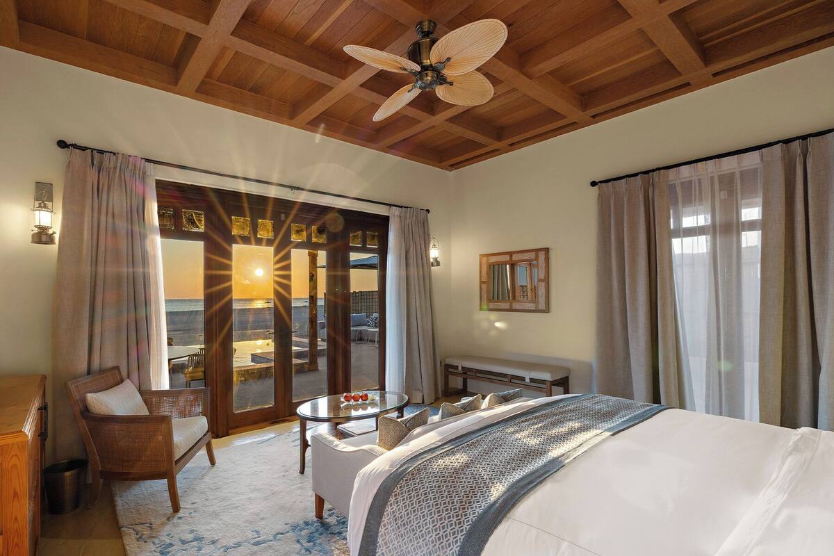 Ein Hotelzimmer mit Bett, einer Sitzecke inklusive Tisch und Stühlen, einem Deckenventilator und Meerblick durch eine Glastür.