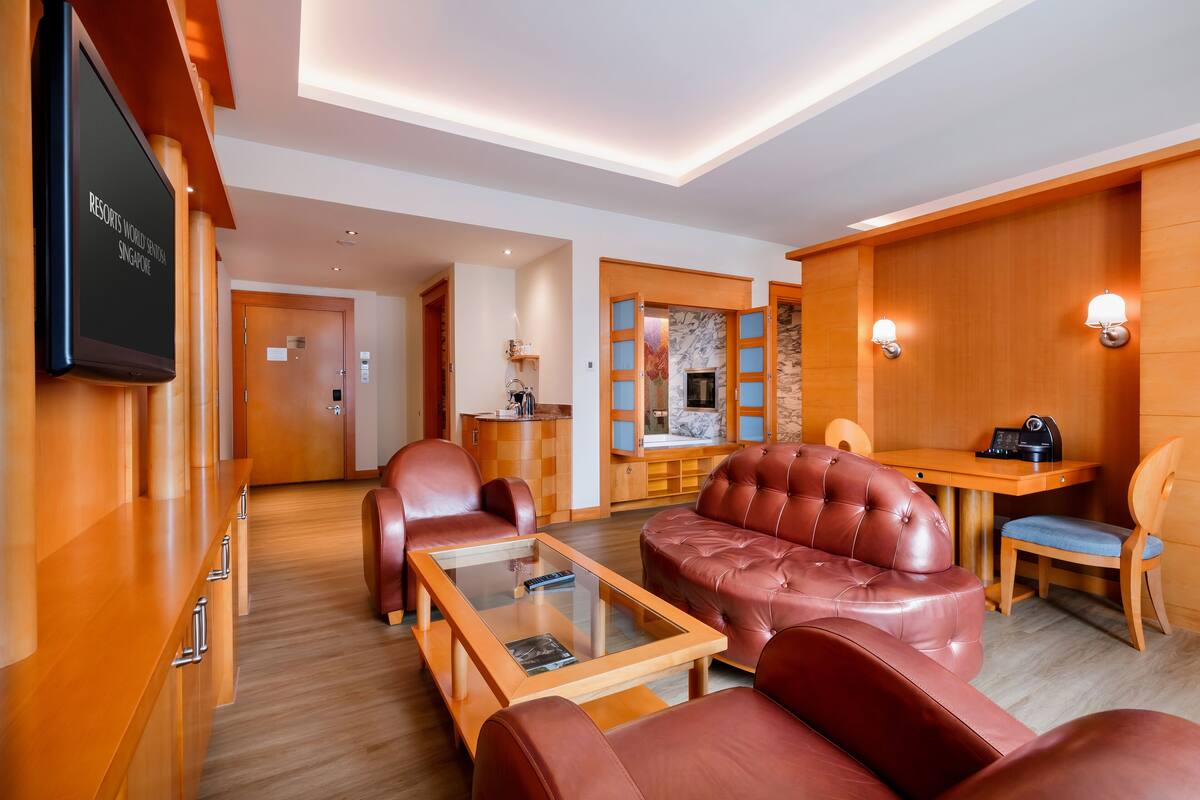 Un hall d’hôtel moderne avec des fauteuils en cuir, une table basse en verre, un comptoir d’accueil en bois et un téléviseur diffusant des images de « RESORTS WORLD SINGAPORE ».