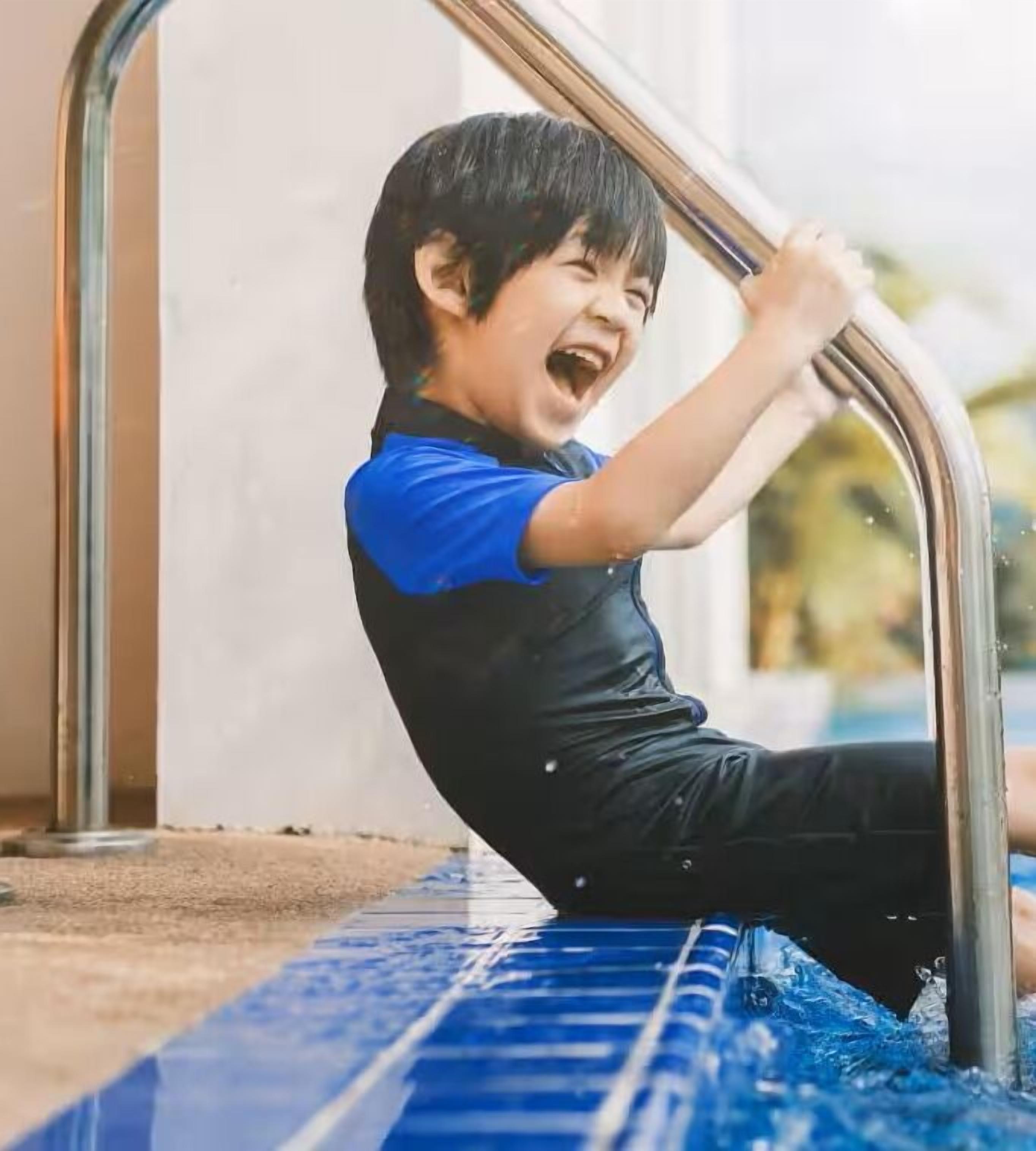 Un enfant est assis sur une surface carrelée bleue à l’extérieur et s’accroche à une rambarde en métal.