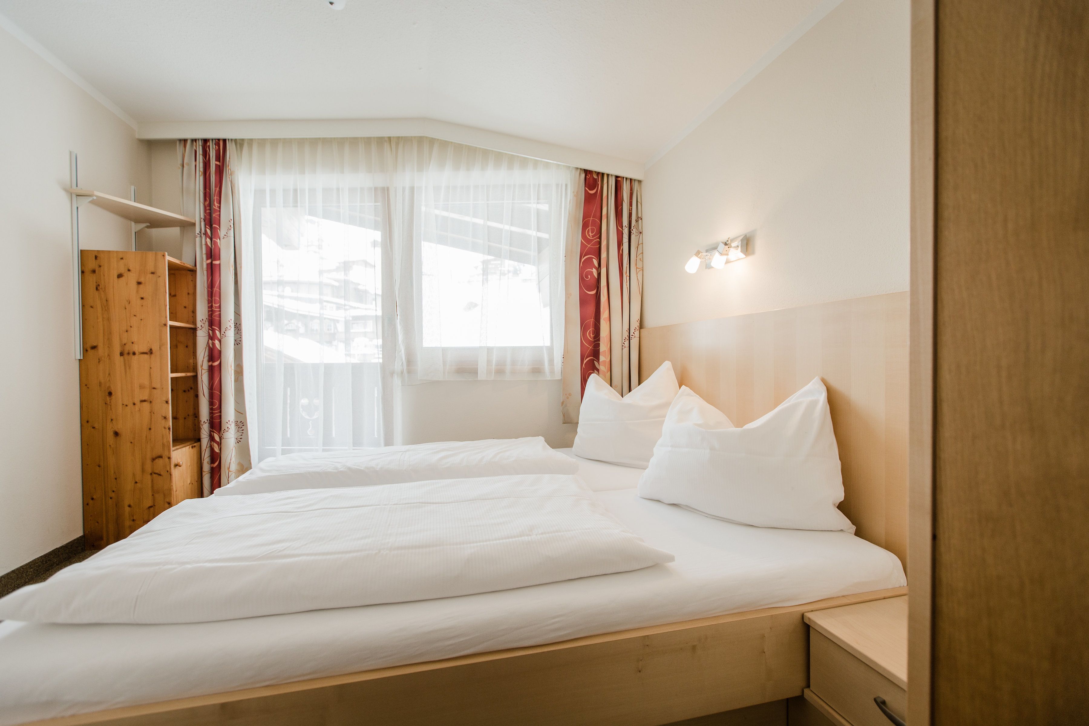 A hotel room with a large bed, wooden headboard, a nightstand, a wooden wardrobe, and a window with sheer curtains.
