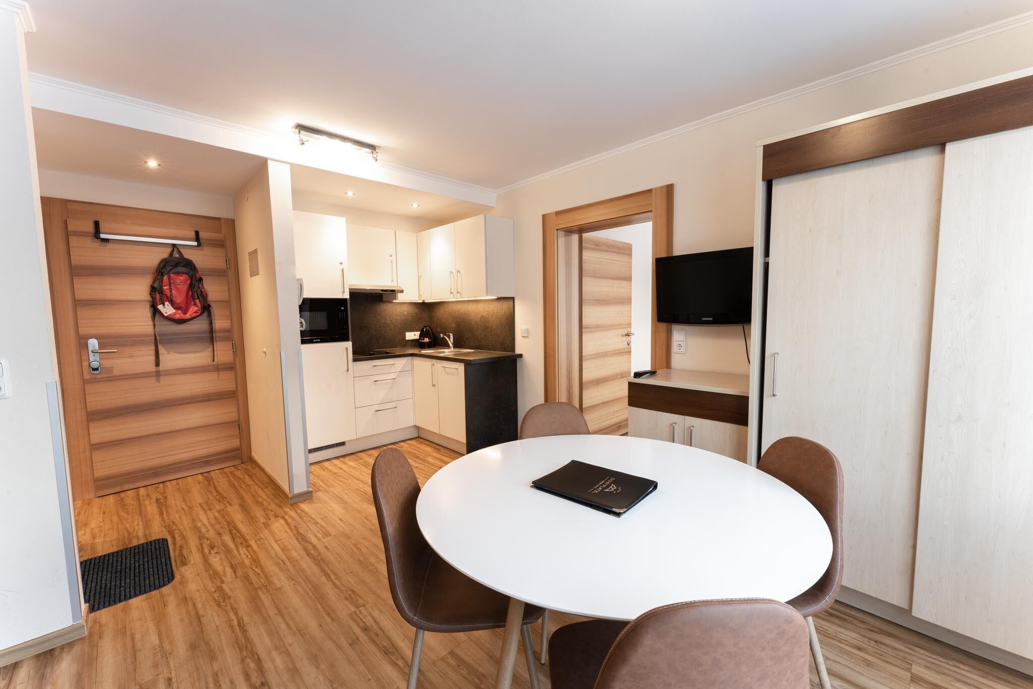 Een moderne keuken met witte kasten, een zwart aanrecht en een magnetron. Er is een eethoek met een witte tafel en bruine stoelen. Een rode rugzak hangt aan de deur.