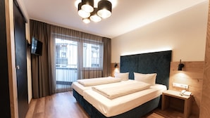Habitación de hotel con una cama grande, un televisor, una mesita de noche y vistas a la ciudad a través del ventanal.