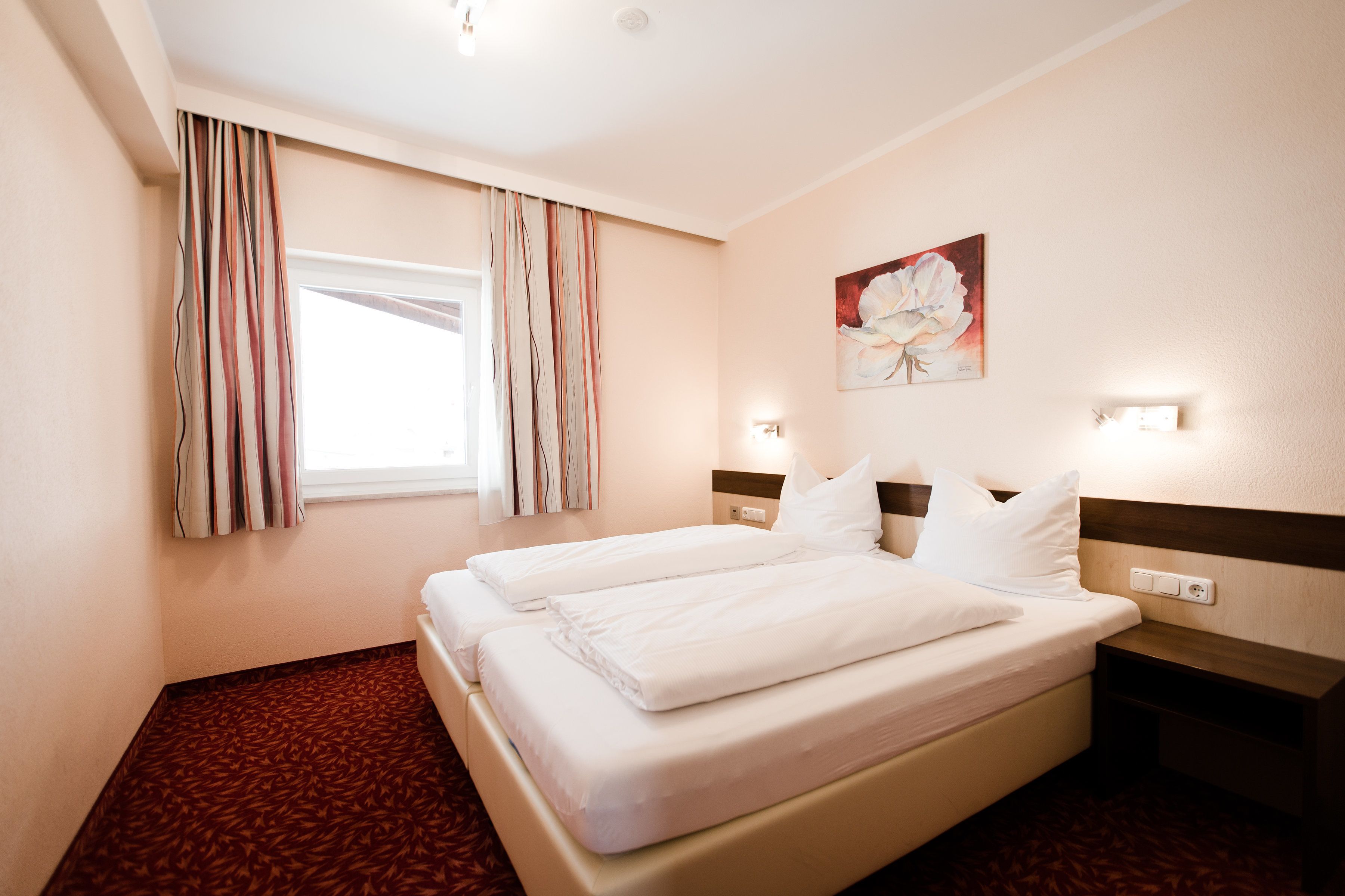 A hotel room with a large bed, a nightstand, a wall-mounted light, and a window with curtains.