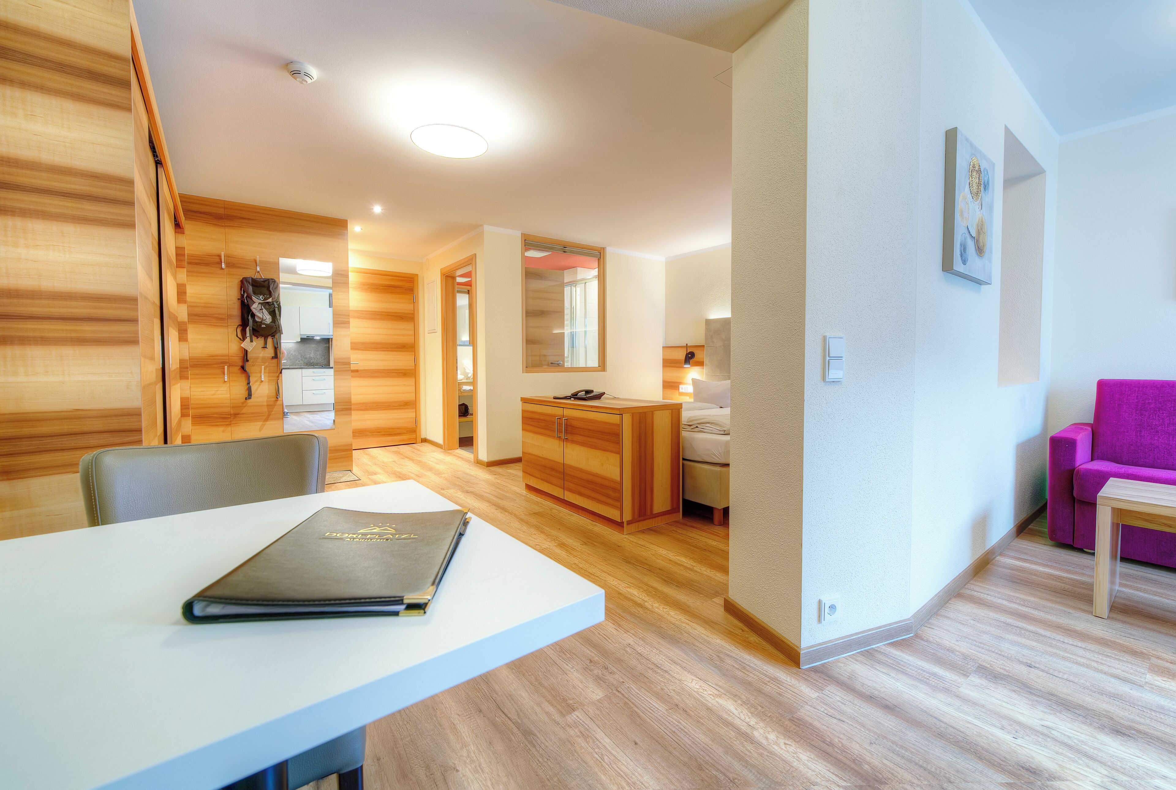 Une chambre d’hôtel moderne avec un plancher en bois, un bureau blanc sur lequel se trouve un livre, un lit, une salle de bain et une chaise rose.