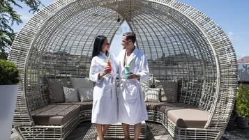 A couple in white robes standing in a circular wicker lounge area with cushioned seating and a hanging light fixture.