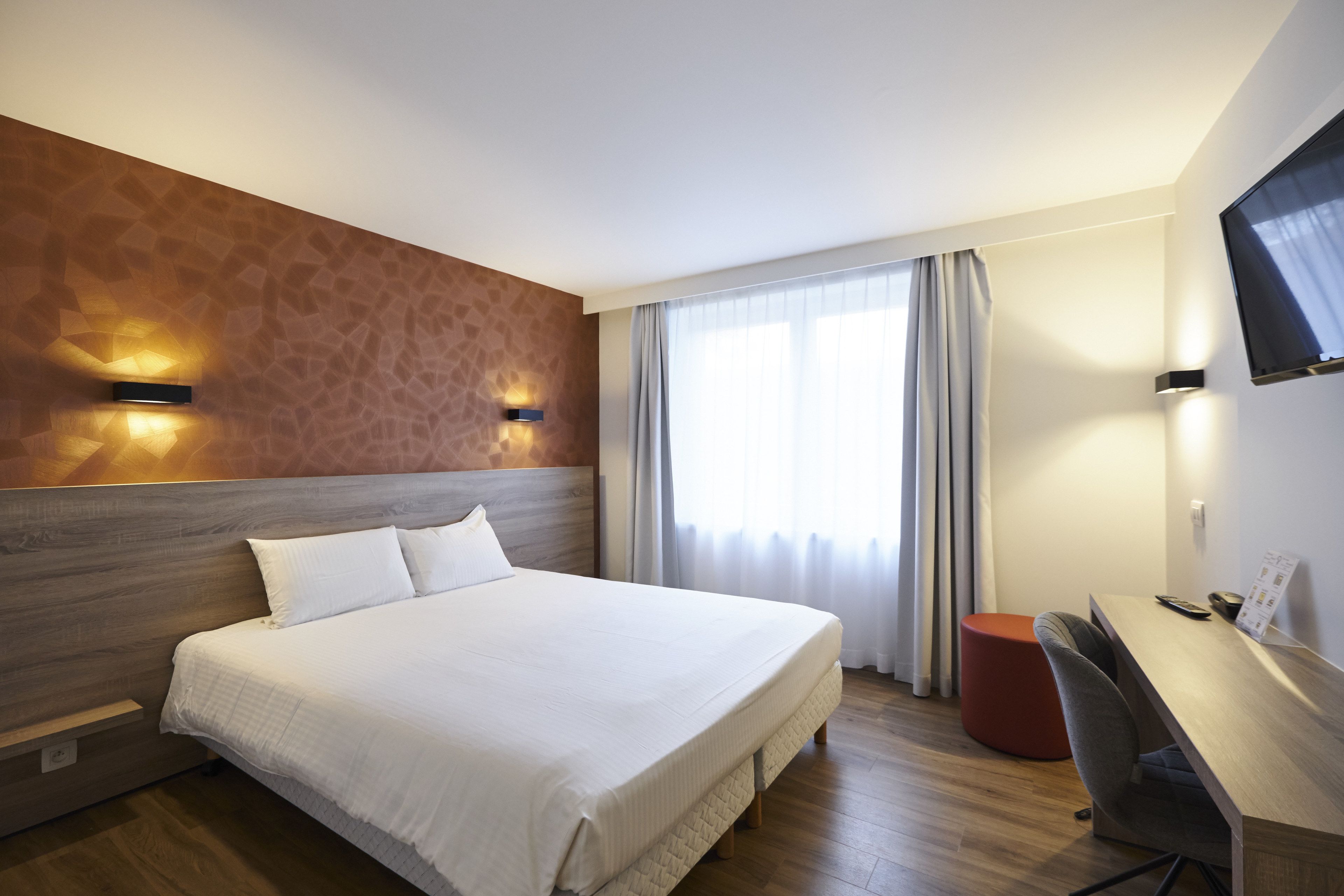 Ein Hotelzimmer mit einem großen Bett, einem Schreibtisch mit Stuhl, einem Fernseher und einem Fenster mit Vorhängen.