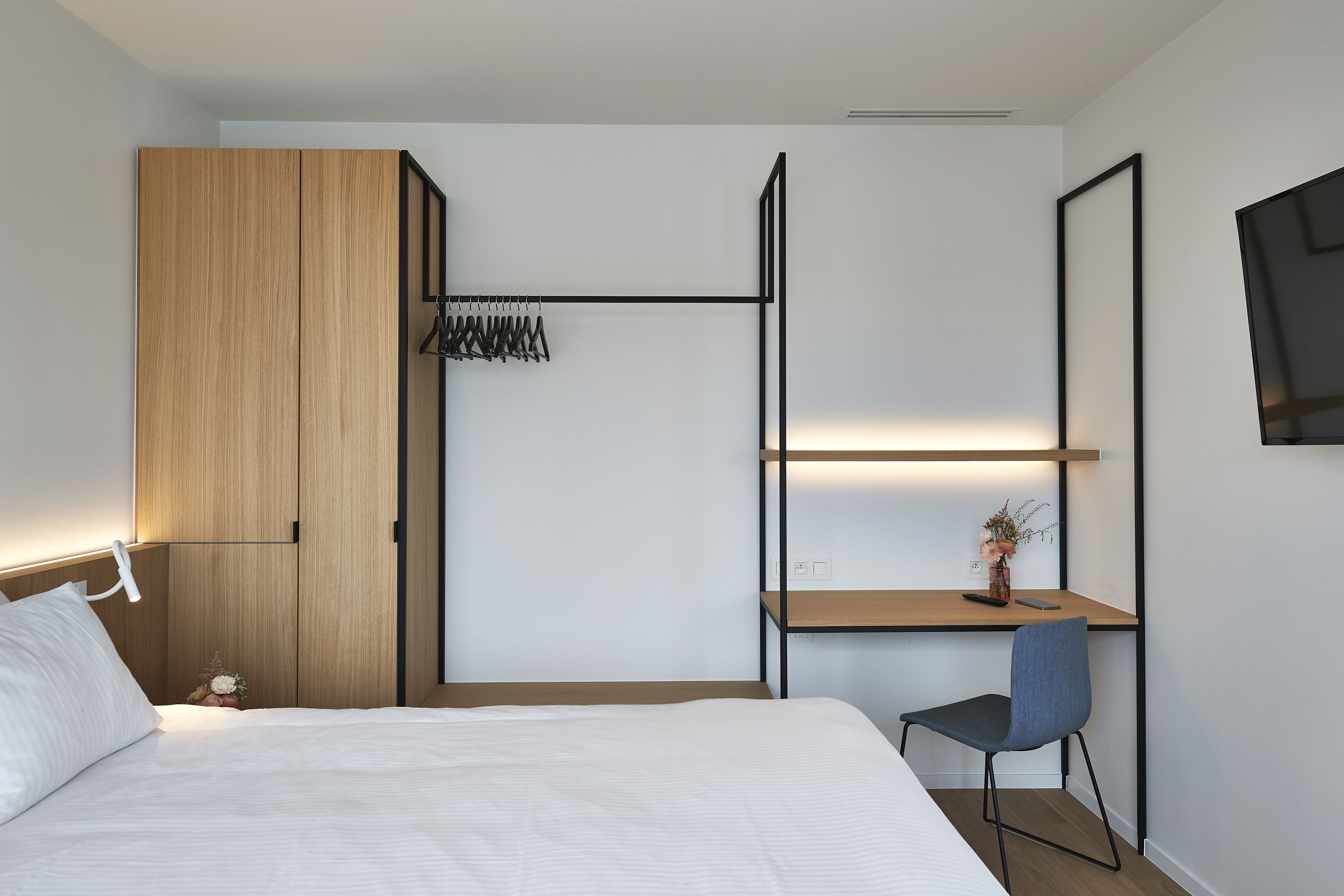 Ein modernes Hotelzimmer mit einem Bett, einem Schreibtisch mit Stuhl, einem Kleiderschrank und einem an der Wand befestigten Fernseher.