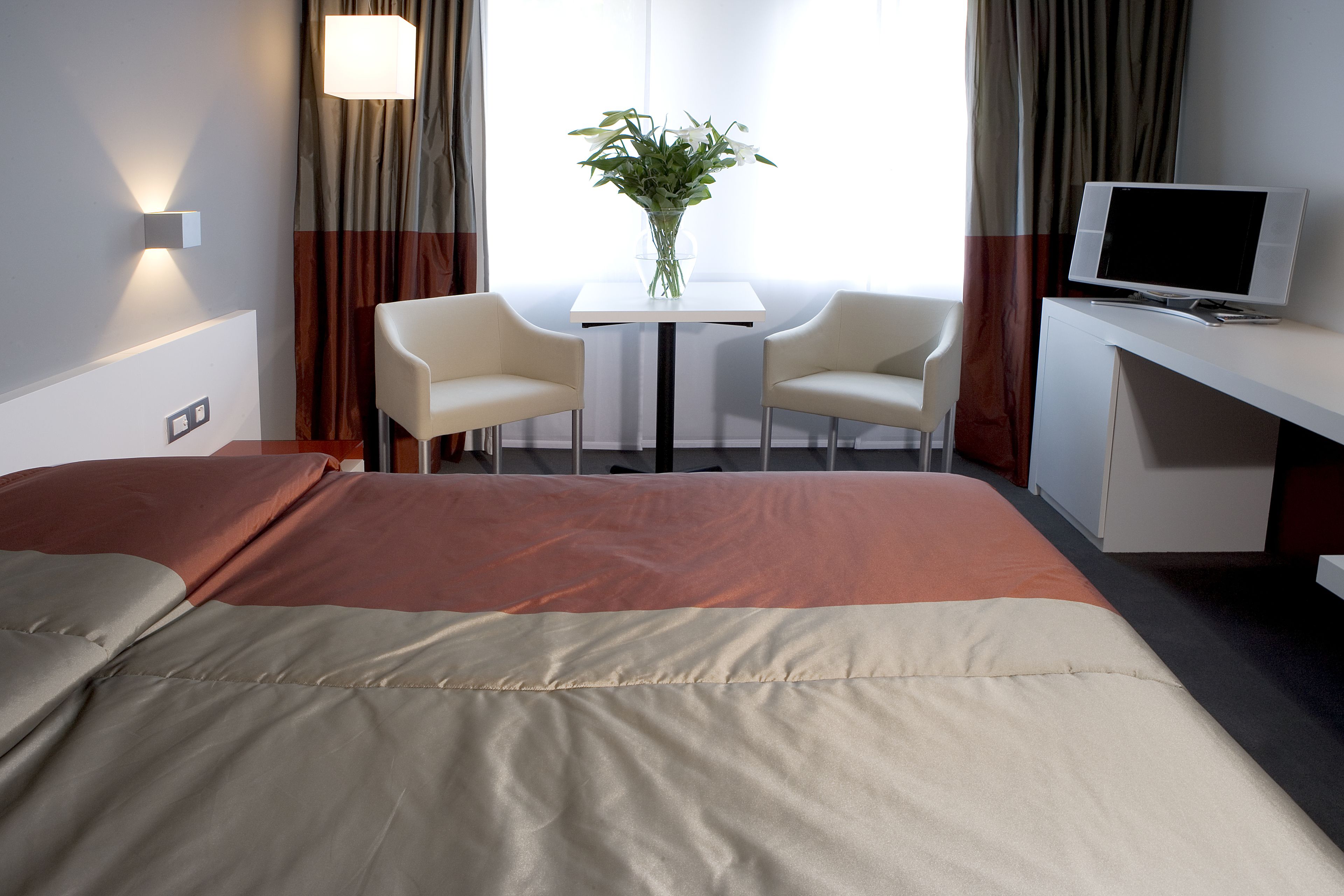 Et hotelværelse med en seng, et skrivebord med tv, to stole, et lille bord og en vase med blomster.