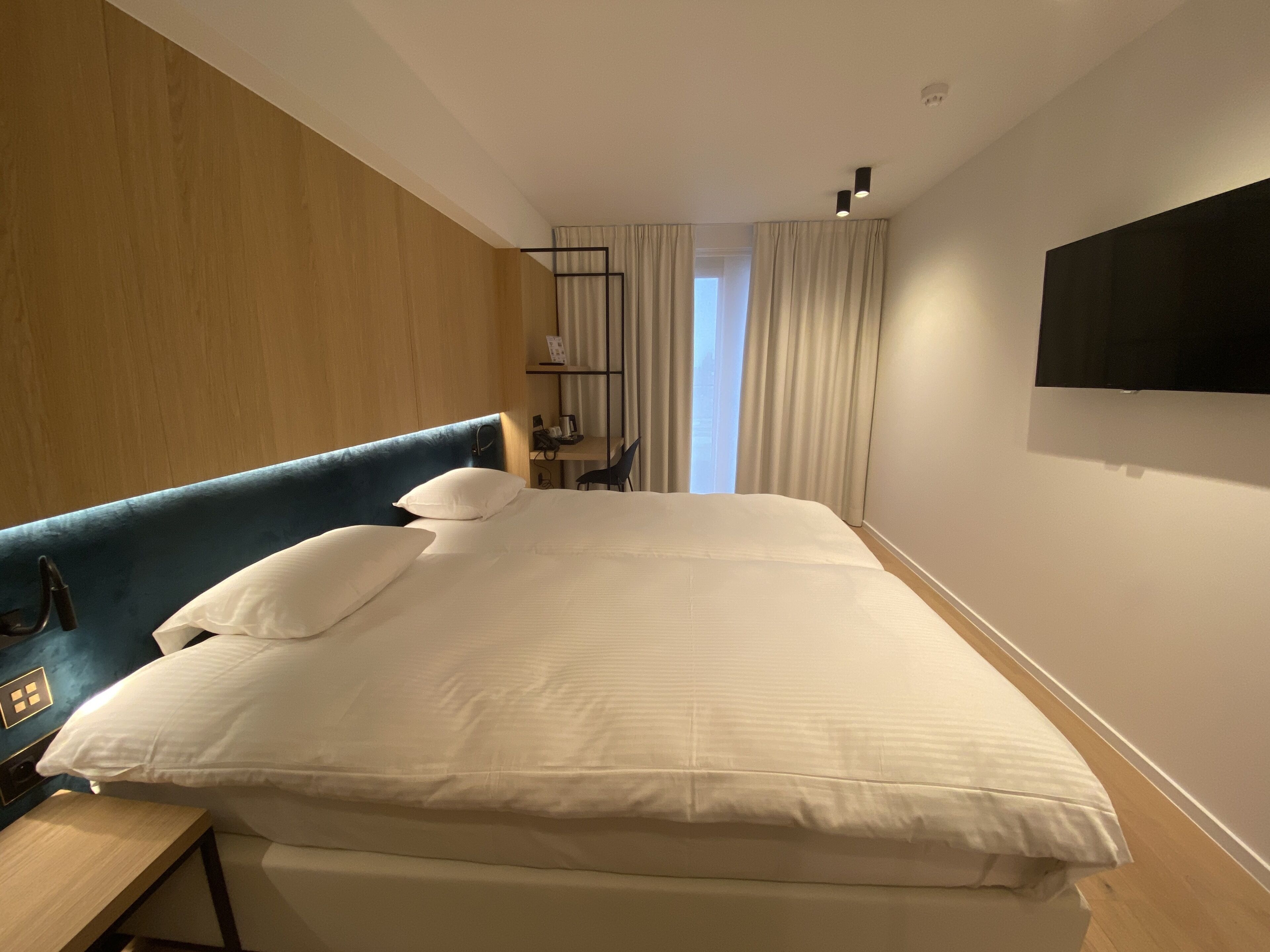 A hotel room with a large bed, a desk, a chair, a TV mounted on the wall, and a window with curtains.