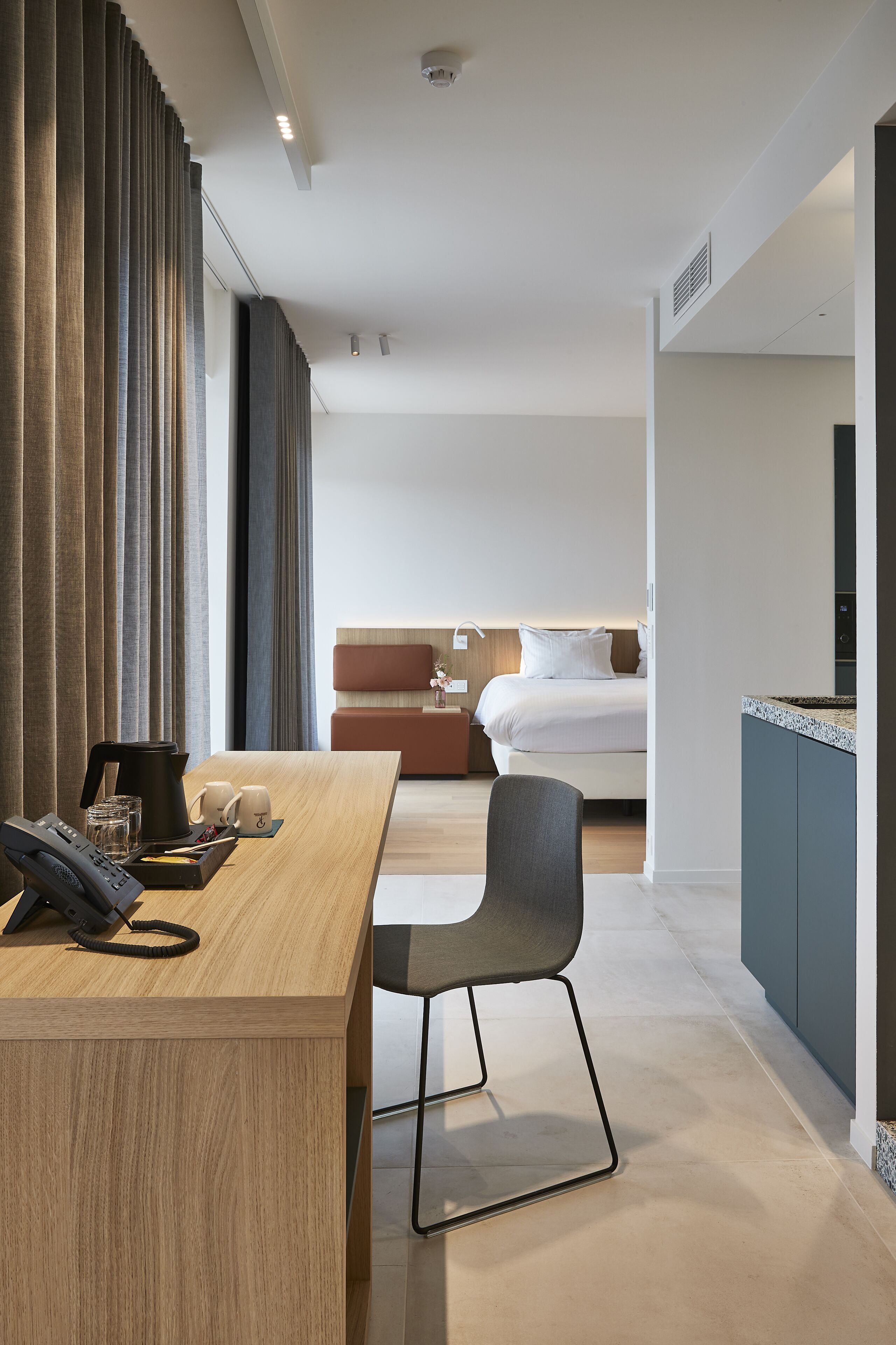 Habitación de hotel con un escritorio de madera, un teléfono y una silla. Se ve una cama con cabecera y almohadas, y una mesita de noche con una lámpara.