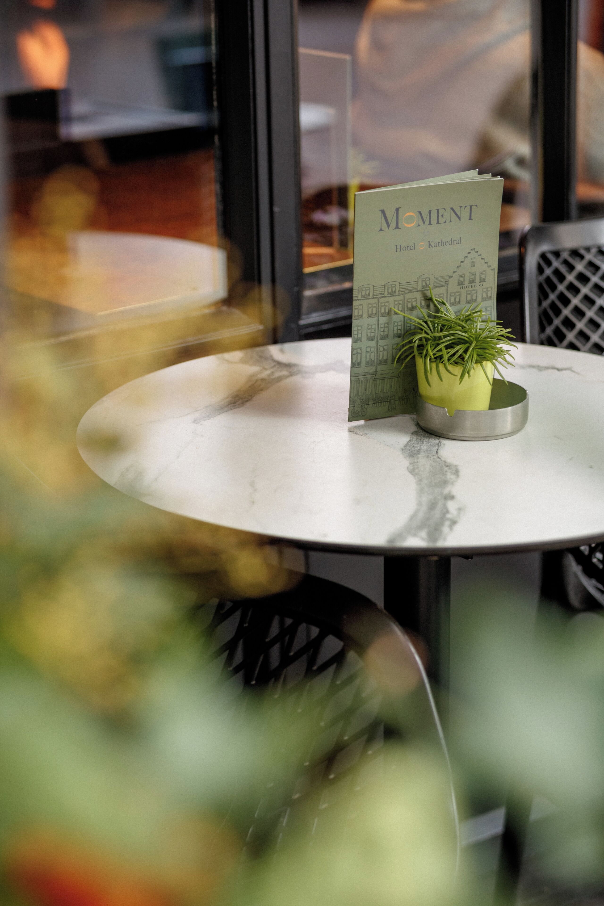 Une table ronde avec un présentoir à menus, une plante en pot et un menu intitulé « Moment Hotel Kathedraal ».