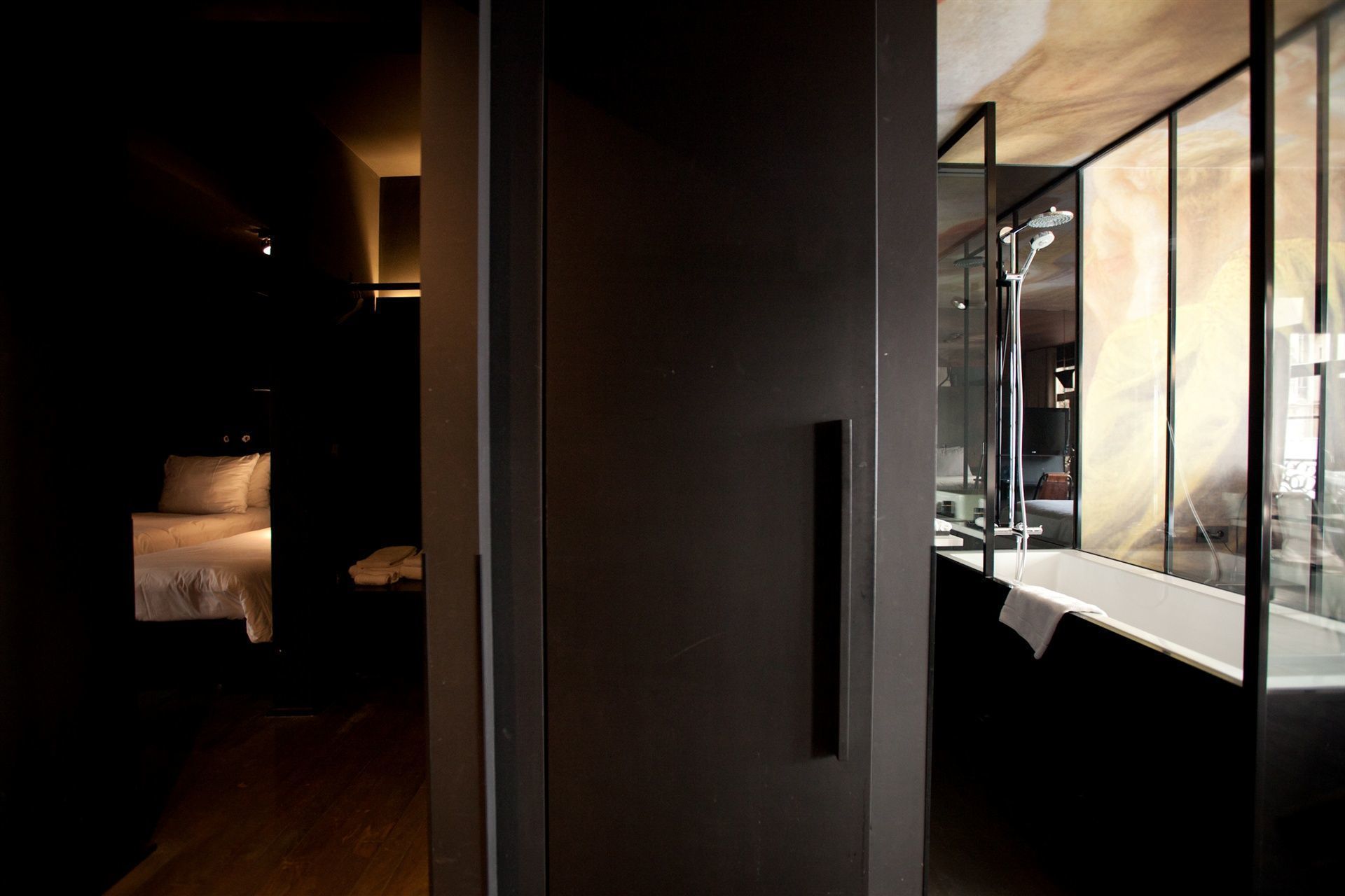 A dimly lit hallway with a bed and a window overlooking a room with a bathtub.