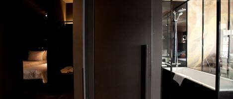 A dimly lit hallway with a bed and a window overlooking a room with a bathtub.