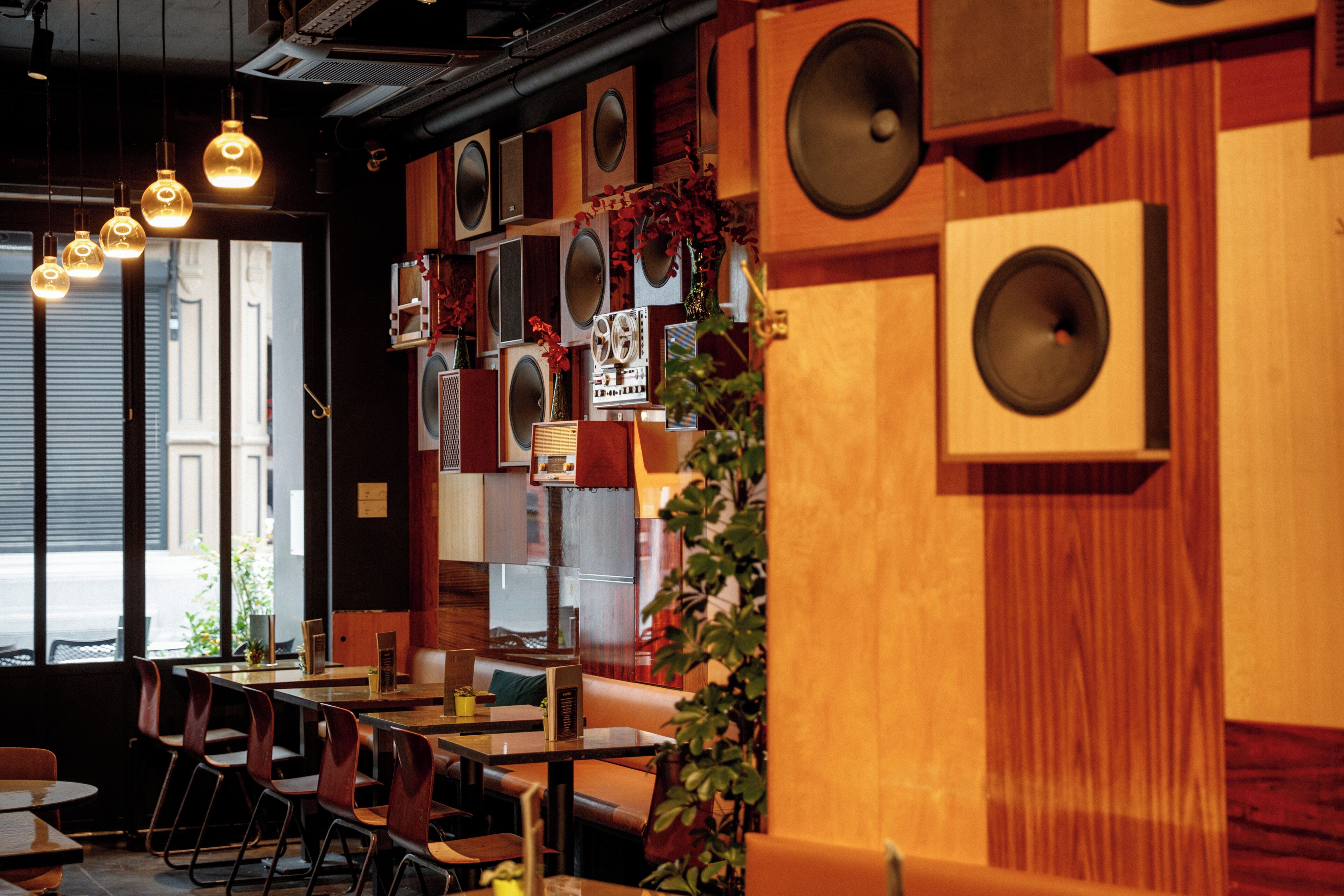 A restaurant interior with multiple tables and chairs, wooden accents, and wall-mounted speakers.