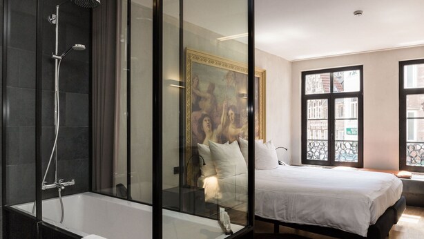A modern bathroom with a glass-enclosed shower, a large bed with white bedding, and a view of the city through large windows. - HotelO Kathedral Antwerp (Antwerp)